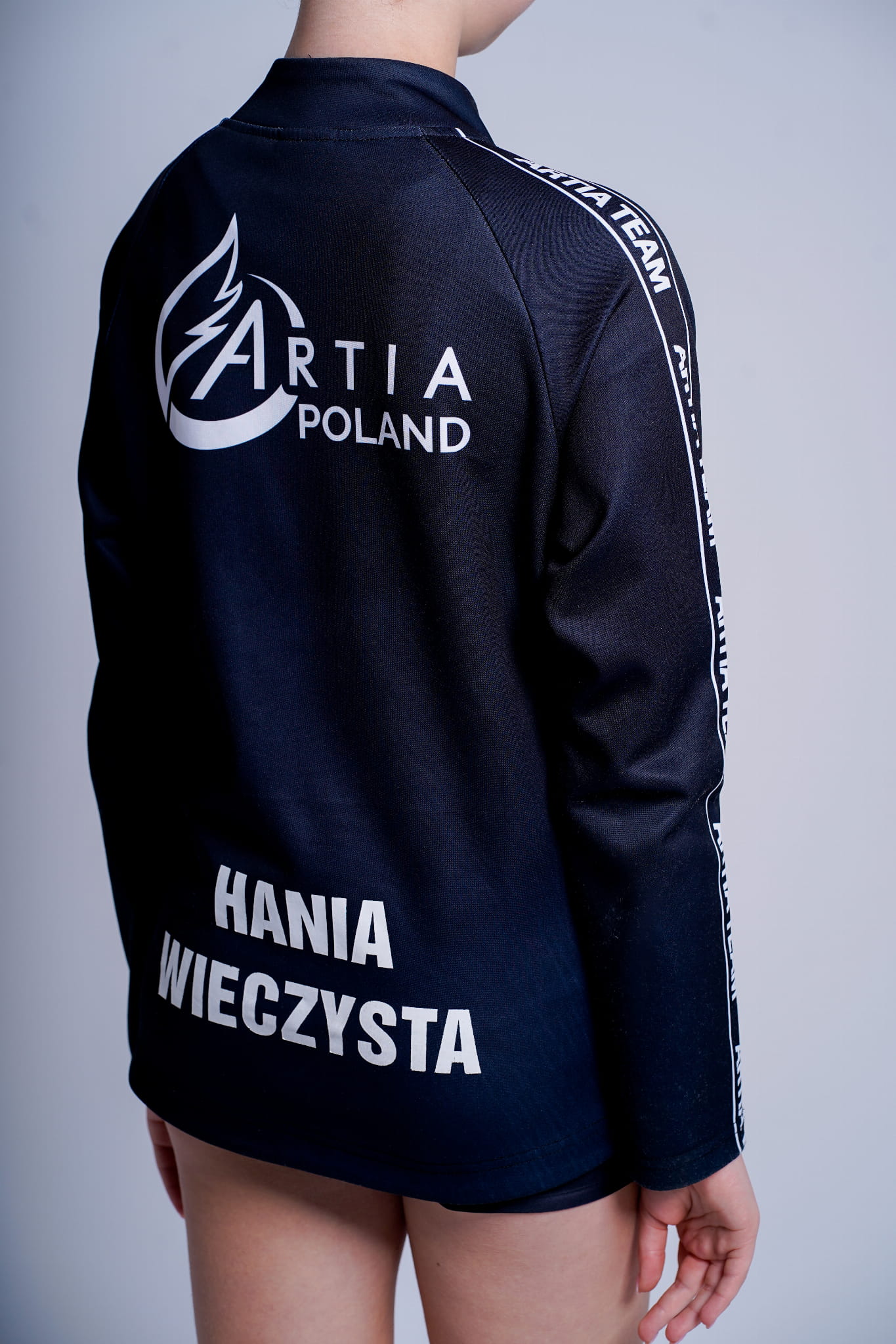 czarna dresowa bluza rozpinana  z białymi napisami artia team na boku oraz logiem Artia Team na plecach.  Na dole bluzy w okolicach bioder napis personalizowany. prezentowane na modelce. widok z tyłu.