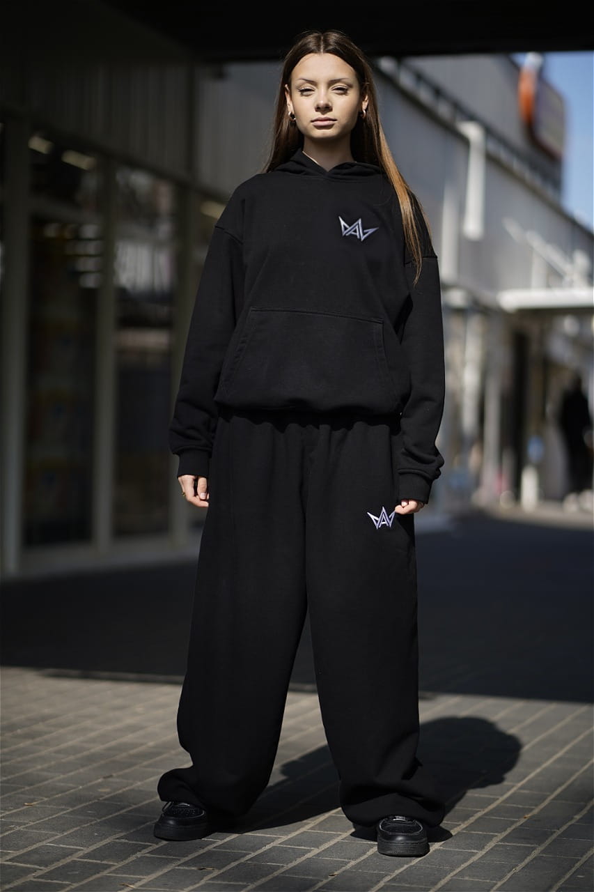 czarna dresowa bluza z kapturem.  z białymi haftowanym znaczkiem artia wear na piersi.  prezentowane na modelce. widok z przodu całej modelki.